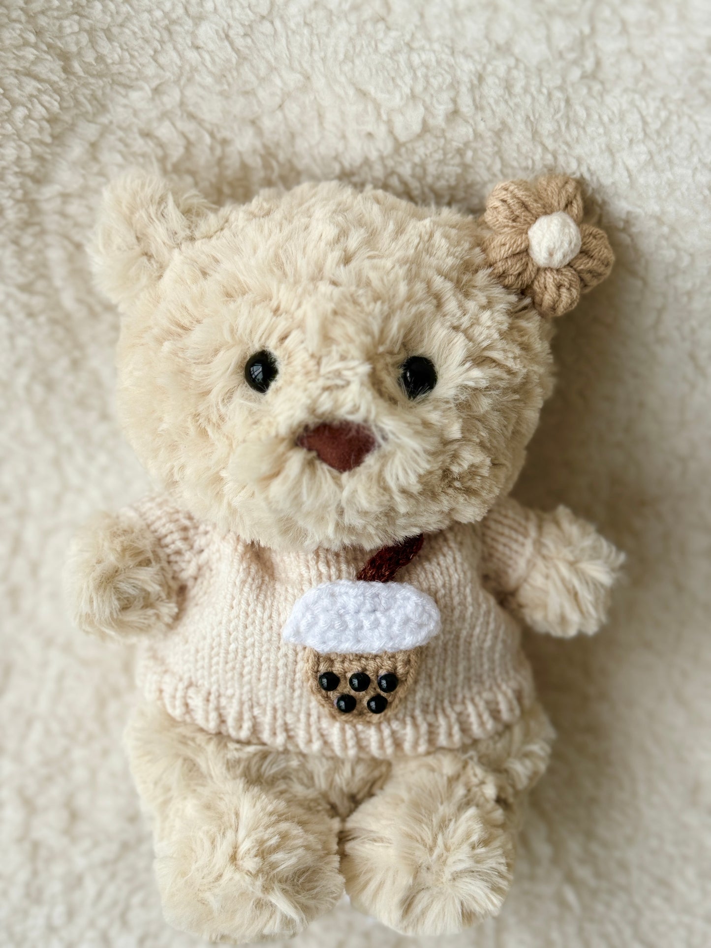 Top-down view of Jellycat Bartholomew Junior in handmade boba tea sweater and crochet flower clip, laid on fluffy background