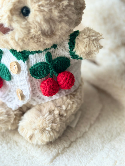 Close-up detail of a handmade cherry cardigan for Jellycat Bart Junior, showing green crochet leaves and bright red cherry appliqué on a white plush sweater.