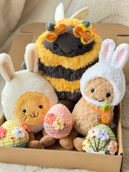 Close-up of three Jellycat plush toys styled for Easter with crochet accessories, including a bunny-hooded plush, a bee plush with butterfly clips, and three pastel crochet Easter eggs decorated with flowers, arranged neatly in a kraft box