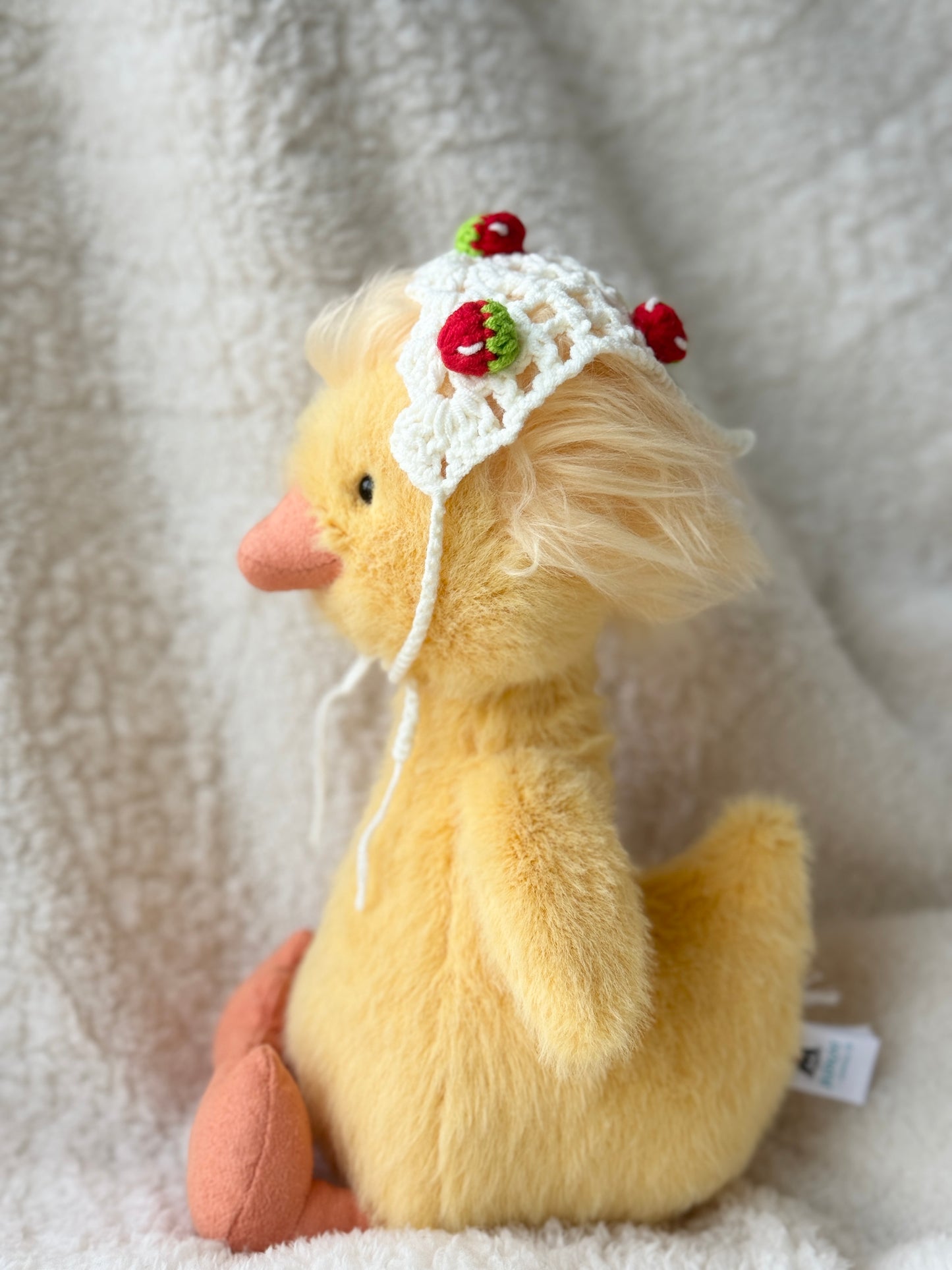 Side view of Jellycat Dorit Duck wearing a handmade crochet strawberry bonnet, a cute plush accessory with cottagecore style and soft cream crochet texture.